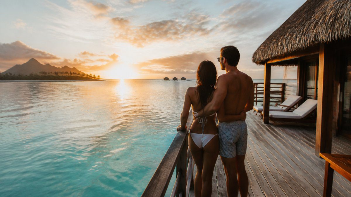 A newlywed couple in swimwear standing on the wooden deck of an overwater bungalow at sunset, turquoise lagoon water below, a tropical island silhouetted in the background